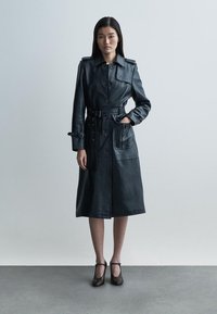 Fekete bőr trench coat öves derékrésszel, vállpántokkal és nagy első zsebekkel. Simább textúra és térdig érő kialakítás.