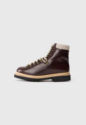 Donkerbruin leren enkelboot met beige veters, metalen oogjes, tan zool, zwarte loopvlak en lichte wollen kraagvoering op een witte achtergrond.