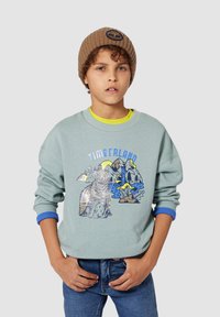 Felpa azzurra chiara con illustrazione di un orso e un paesaggio montano, con polsini a coste blu e colletto rotondo. Berretto marrone con logo.