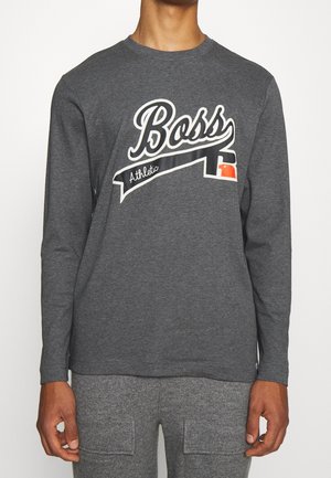 Mężczyzna w ciemnoszarej koszulce z długim rękawem z napisem "Boss Athletic" oraz małą, czerwoną i czarną grafiką na przodzie.