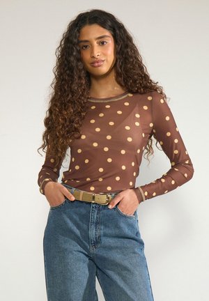 Ung kvinde med langt krøllet hår iført en brun polkaprikket langærmet top, blå jeans og et beige bælte, hænderne i lommerne, neutralt baggrund.