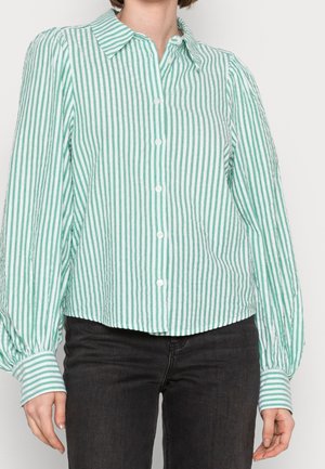 Personne portant une chemise boutonnée à manches longues à rayures verticales vertes et blanches avec des poignets plissés et un pantalon noir.