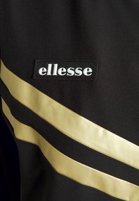 Ropa negra con dos franjas doradas anchas y un logo blanco de "ellesse" en una superficie de tejido texturizado.