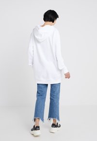 Felpa bianca con cappuccio e vestibilità ampia, abbinata a jeans blu con orlo sfrangiato e sneakers nere chunky, vista da dietro.