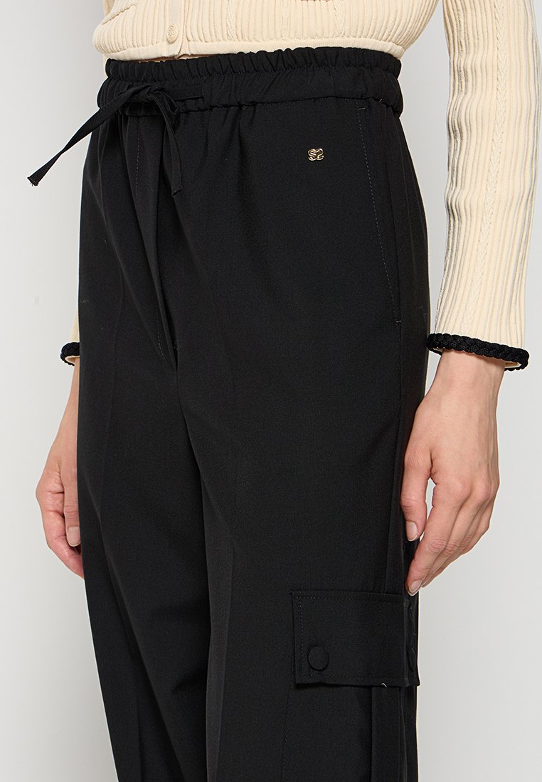 Zwarte cargobroek met een elastische tailleband, trekkoord, zijzakken en een knoopzakdetail. Gladde textuur met een gouden logoaccent.