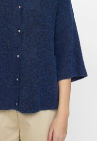 Mørkeblå strikket cardigan med korte ærmer, struktureret stof, perlelignende knapper og en løs pasform, parret med beige bukser.