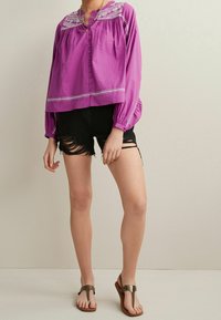 Lilla langærmet bluse med knaplukning og broderet halsudskæring; kombineret med slidte sorte shorts og brune sandaler.