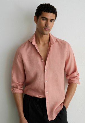 Mann mit lockigem schwarzen Haar, der ein aufgeknöpftes rosa Langarmhemd und schwarze Hose trägt, steht vor einer einfachen weißen Wand.
