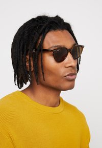 Ochelari de soare cu imitate tortoise, lentile de culoare brună și accente metalice. Modelul poartă un pulover tricotat galben cu decolteu rotund.