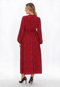 Largo vestido floral rojo con mangas largas y cintura ajustada, con un lazo en la espalda y una falda fluida. Combinado con tacones altos negros.
