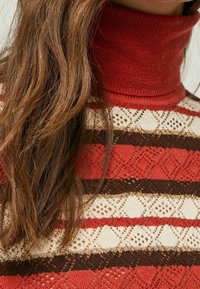 Röd och krämfärgad stickad tröja med rullad polokrage, med horisontella ränder av spets och enfärgade nyanser i brunt, orange och kräm.