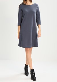 Harmaa, polvipituinen mekko, jossa on kolme neljänneksen hihteä, pyöreä kaula-aukko ja rento istuvuus. Yhdistetty mustiin nilkkureihin. Yksinkertainen ja sileä rakenne.