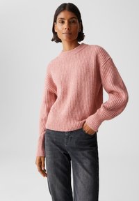 Frau steht mit der Hand in der Tasche, trägt einen gerippten pinken Pullover und dunkle graue Jeans vor einem schlichten Hintergrund.