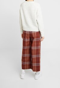 Hellgrauer Pullover, weite karierte Hose in Rot und Braun, weiße Sneakers. Der Pullover hat einen Rundhalsausschnitt; die Hose weist ein Karomuster auf.