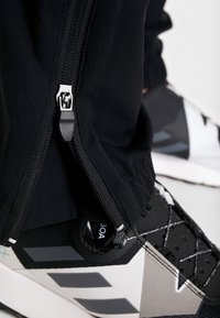 Pantalones negros con cremallera y acabado texturizado, combinados con zapatos deportivos negros y grises que presentan un diseño estampado y un sistema de cordones único.