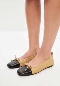 Ballerine con tomaia in pelle beige e punta in pelle nera. Presentano una suola piatta e un dettaglio a fiocco nella parte anteriore.