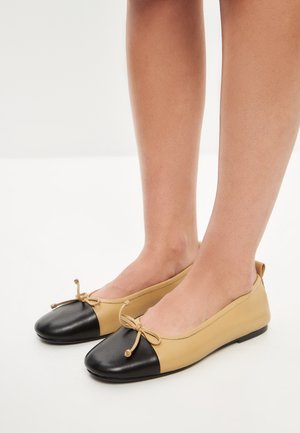Ballet flats met een beige leren bovenwerk en een zwart leren neuskap. Heeft een platte zool en een strikdetail aan de voorkant.