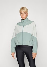 Chaqueta recortada en verde claro y blanco con un alto cuello, puños elásticos y cinturilla. Cuenta con cierre de cremallera y logo de la marca en el pecho.