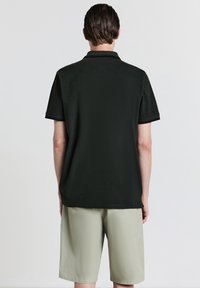 Uomo che indossa una polo nera a maniche corte e pantaloni corti beige chiaro fino al ginocchio, in piedi di spalle su uno sfondo bianco semplice.