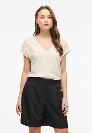 Kobieta o długich brązowych włosach, ubrana w kremowy krótki top z dekoltem w serek i wysokim stanem czarne plisowane szorty, stojąca na białym tle.