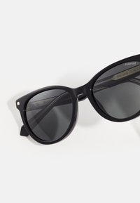 Occhiali da sole in plastica nera con lenti grigie, design angolare e dettagli dorati sulla montatura. Presentano lenti ampie e una struttura resistente.