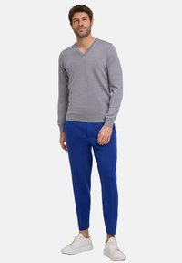 Grauer V-Ausschnitt-Pullover kombiniert mit blauen, schmalen Hosen und weißen Sneakers. Der Pullover hat eine glatte Textur; die Hose weist ausgeprägte Nähte auf.