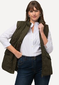 Oljen grøn quiltet vest med framside glidelås og trykknapp lukking, stylet over en hvit krage skjorte. Har sidelommer og en figursydd design.