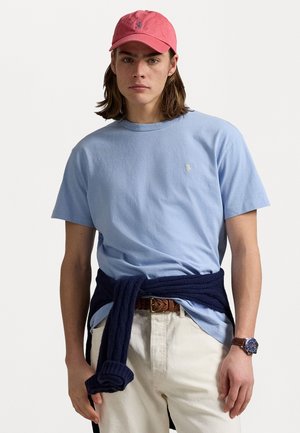 Light blue cotton t-shirt with a small logo, navy sweater tied around the waist, beige pants, and a pink cap. Brown belt with a watch.