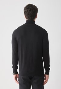 Hombre con cabello rizado oscuro, vistiendo un suéter negro de cuello alto y jeans oscuros, de pie de espaldas contra un fondo blanco.