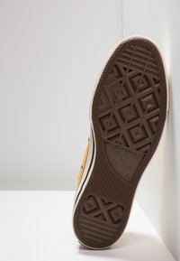 Gule lerret sneakers med en teksturert brun gummisåle med geometrisk mønster, sorte stripeaksenter og et klassisk design.