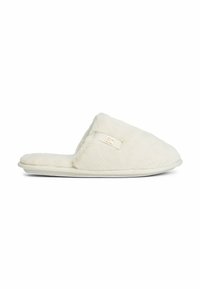 Zachte, crèmekleurige instapslippers met een pluizige textuur en een klein decoratief logo aan de zijkant, vlakke zool voor binnengebruik.