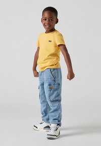 Žluté tričko s krátkým rukávem s logem, světle modré kapsáčové džíny s kapsami a černo-bílé tenisky. Jednoduchý a ležérní outfit.