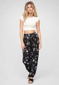 Jeune femme portant un crop top blanc, un pantalon noir à fleurs avec cordon, et des sandales noires, se tenant devant un fond clair.
