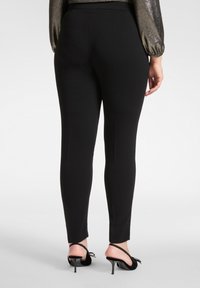 Schwarze, eng anliegende Hosen mit glatter Textur und knöchellangem Design, ergänzt durch Stöckelschuhe mit Schleifen auf den Riemen.