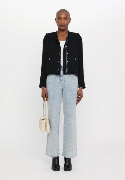 Schwarze cropped Jacke aus strukturiertem Material, mit silbernen Knöpfen und fransigen Kanten, kombiniert mit hellblauen Schlagjeans und schwarzen Stiefeletten.