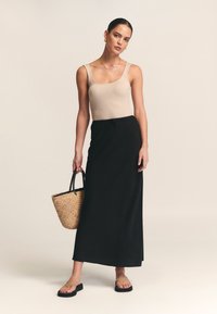 Beiges geripptes Tanktop, schwarzer Maxirock und schwarze Flip-Flops. Das Modell hält eine gewebte Strohtasche mit schwarzen Griffen. Sauberes und minimalistisches Design.