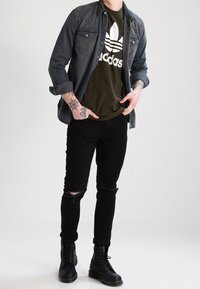 Camisa de mezclilla oscura sobre una camiseta con logo de Adidas en verde oliva, jeans rasgados negros y botas negras con cordones, presentado contra un fondo neutro.