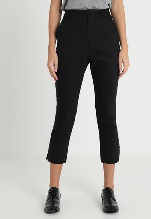 Kobieta w czarnych wąskich spodniach o krótkim kroju z szlufkami na pasek i czarnych eleganckich butach sznurowanych, pokazana od połowy tułowia do stóp na jednolitym tle.