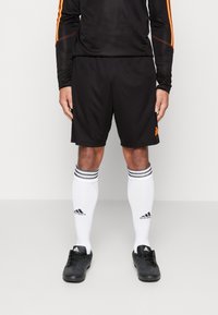 Person som bär svarta sportshorts, en lång svart tröja med orange ränder, vita knähöga Adidas-strumpor och svarta Adidas fotbollsskor.