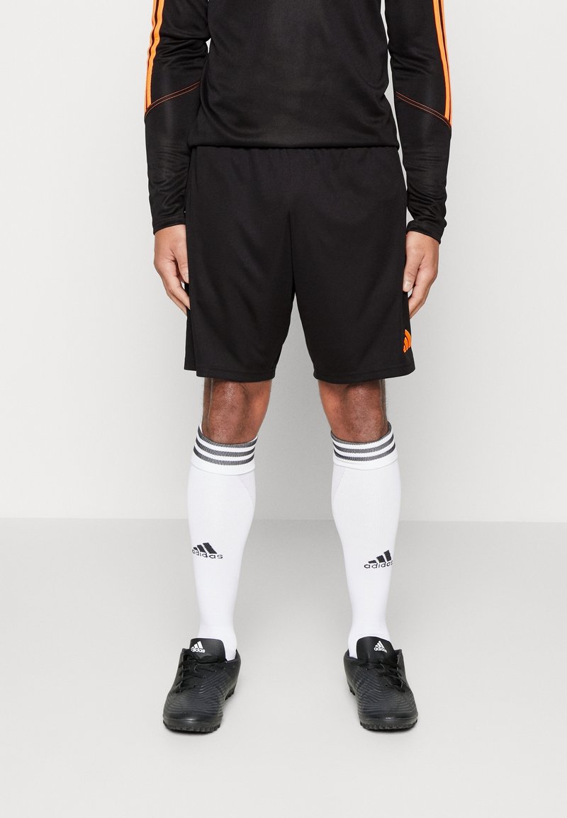 Person som bär svarta sportshorts, en lång svart tröja med orange ränder, vita knähöga Adidas-strumpor och svarta Adidas fotbollsskor.