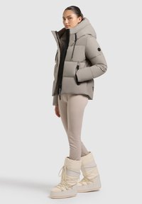 Doudoune beige avec un col haut, des zips noirs et des poches, associée à des leggings beige clair et des bottes de neige blanches.