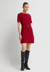 Rode jurk met korte mouwen en een getailleerd design, voorzien van een gouden accent bij de taille, gecombineerd met kniehoge zwarte leren laarzen.