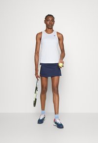 Hvit ermeløs sportstopp, marineblå skjørt, holder en tennisball, holder et racket, iført blå og røde treningssko med lyseblå sokker.