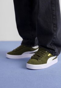 Grønne semskede sneakers med hvite detaljer, rund tå, flat såle og gylden merkevaredetalj, kombinert med mørke denimjeans på en lys blå overflate.