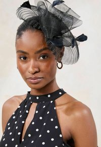 Halterica s crnim i bijelim točkicama s otvorom za ključ; sadrži crnu mrežastu fascinator s pernatim naglascima. Glatka tekstura tkanine.