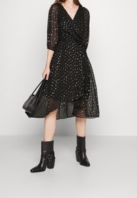 Robe portefeuille noire à paillettes dorées, manches longues transparentes, taille nouée, associée à des bottines noires à bride et boucle.