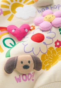 Tissu crème avec des appliqués brodés colorés : un chien marron, un cœur rose, une fleur jaune et un soleil, avec les éléments de texte "hop hop" et "WOOF!"