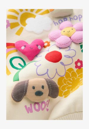 Tissu crème avec des appliqués brodés colorés : un chien marron, un cœur rose, une fleur jaune et un soleil, avec les éléments de texte "hop hop" et "WOOF!"