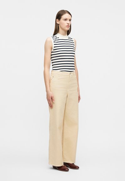 Γυναίκα που φοράει μπεζ παντελόνι φαρδύ στο πόδι, αμάνικη κοντομάνικη μπλούζα  με άσπρες και μαύρες ρίγες και καφέ παπούτσια, στέκεται μπροστά σε ένα απλό λευκό φόντο.