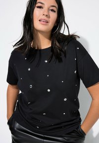Camiseta negra de manga corta en tejido suave, adornada con embellissements circulares metálicos de diferentes tamaños. Combinada con pantalones negros.